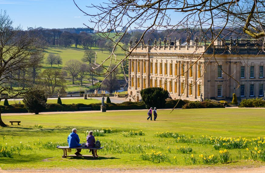 An enormous manor house stands in landscaped grounds with many trees; two visitors are sitting on a bench in the foreground, while two others are walking between them and the house.