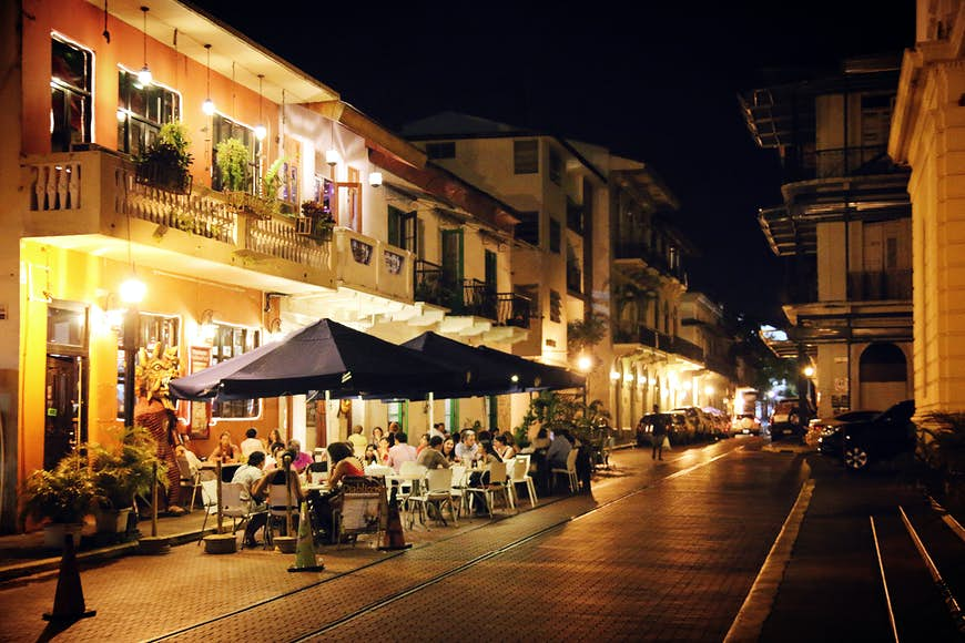 Groups of people sit at tables installed with large umbrellas outside of a restaurant next to cobbled stone streets in the historic Casco Viejo