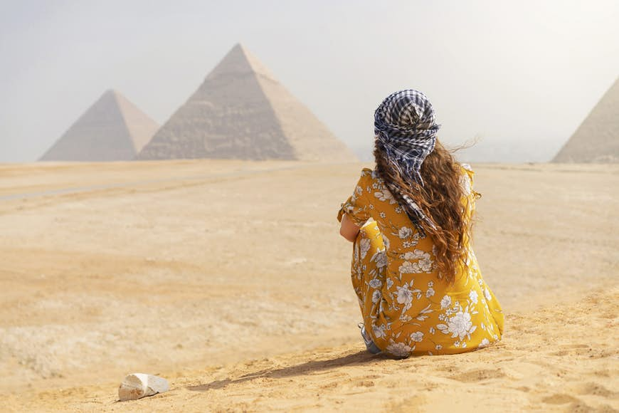 A female tourist sitting on a sand dune and looking at the Pyramids of Giza.