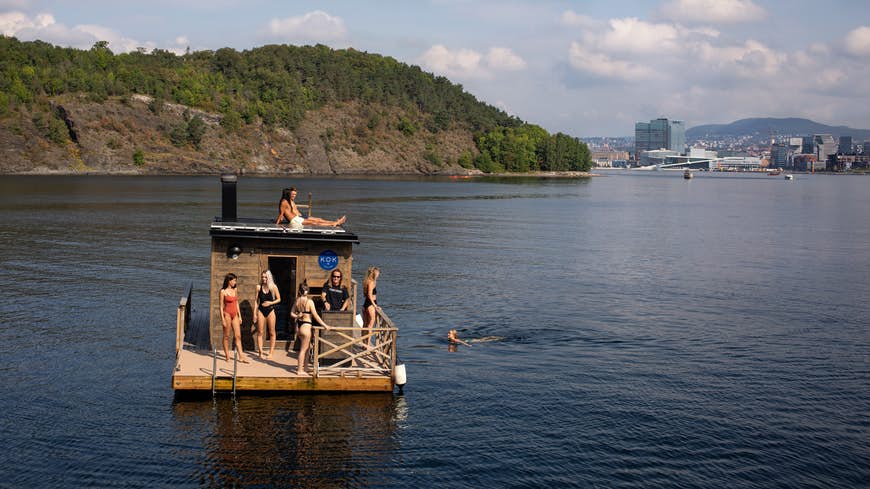 A group of people use the KOK floating sauna on the fjord next to Oslo. One is swimming, another sunbathing on the roof, and others stood on the deck. The driver is wearing a black t-shirt.  