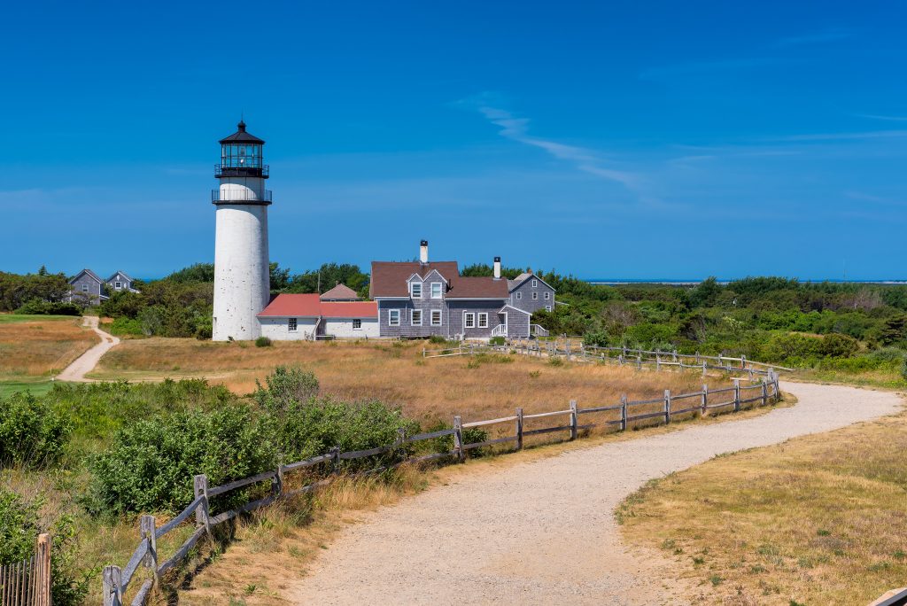 A path leading to a tall white lighthouse attached to a small house, underneath a bright blue sky.