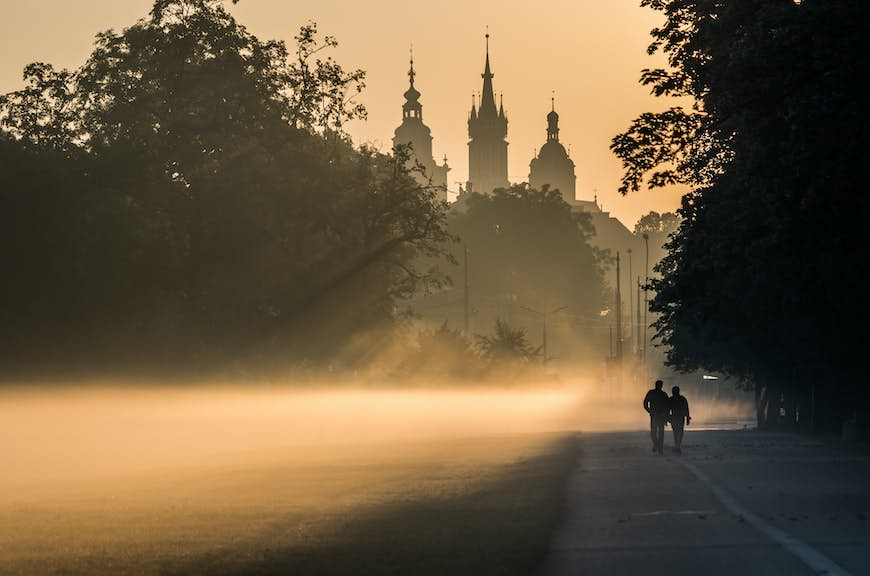 Two people in silhouette walk through a misty park with a large castle building in the distance