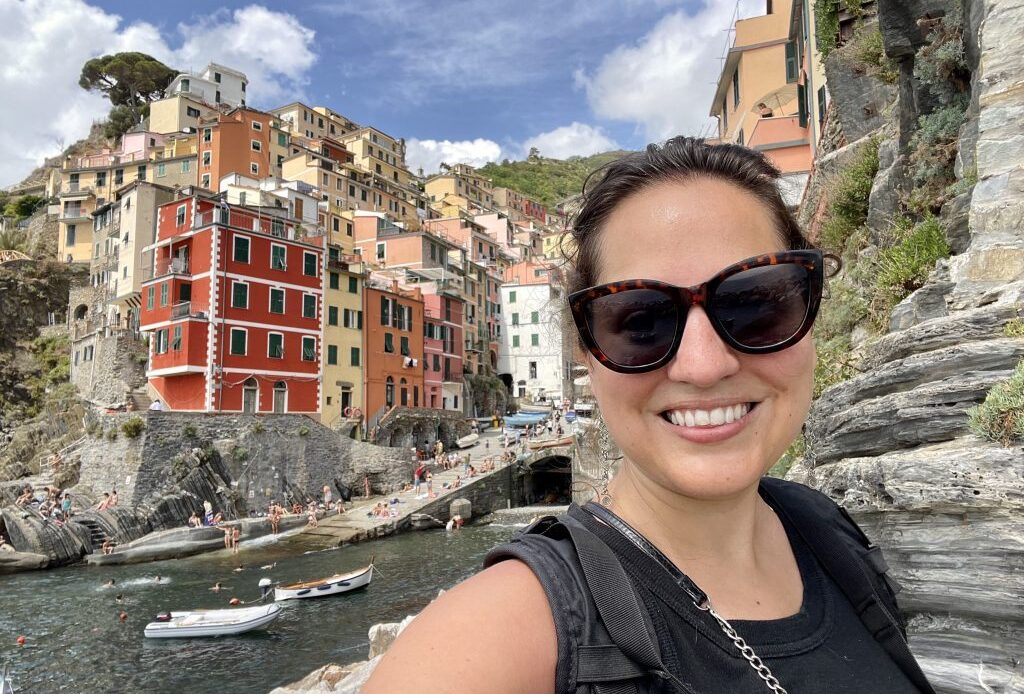 Kate taking a smiling selfie in sunglasses in front of the steep colorful buildings of Vernazza in Cinque Terre, all built into a rocky cliff.