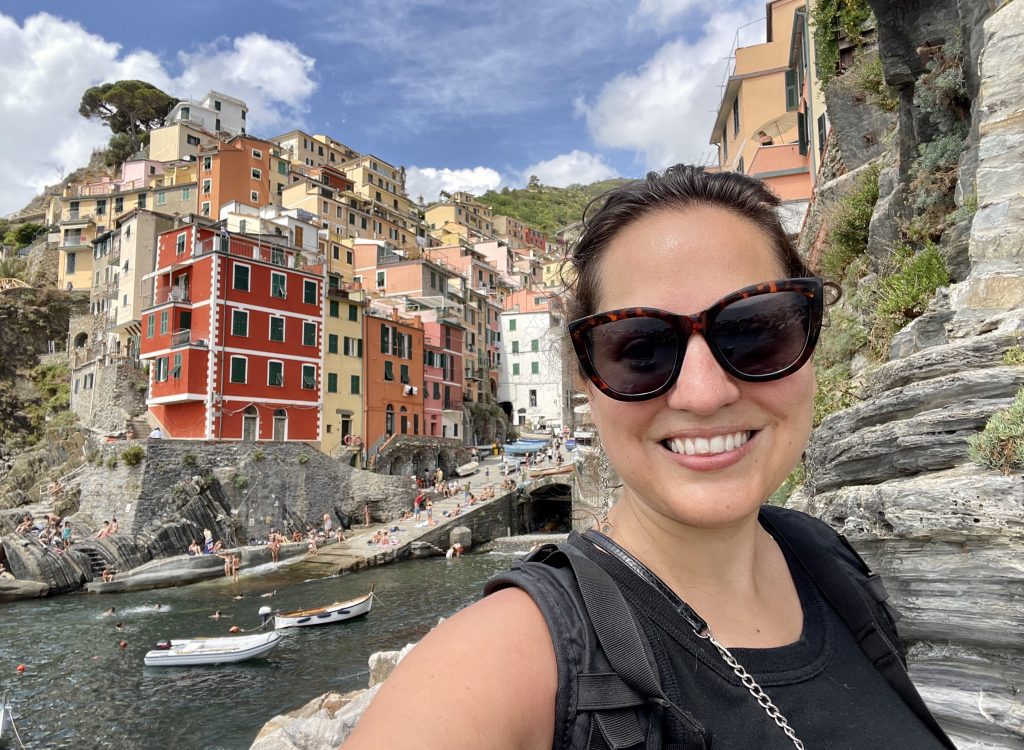 Kate taking a smiling selfie in sunglasses in front of the steep colorful buildings of Vernazza in Cinque Terre, all built into a rocky cliff.
