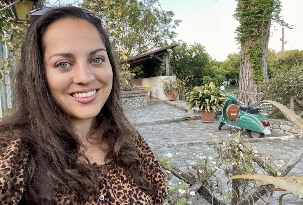 Kate taking a smiling selfie in a leopard-print shirt, behind her is a rural guesthouse with gardens and a turquoise Vespa.