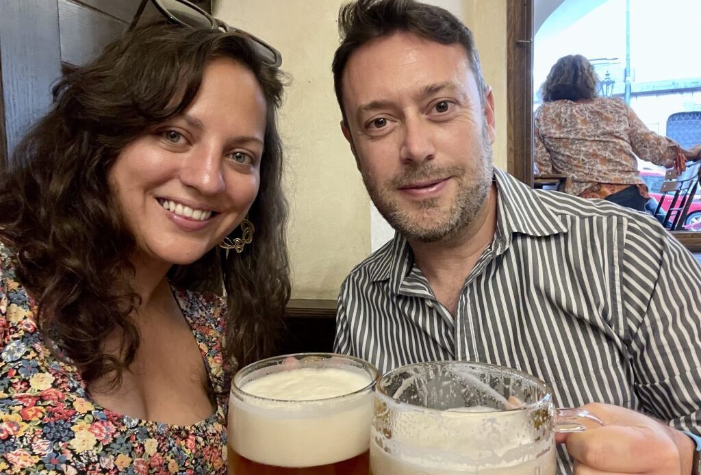 Kate and Charlie hold us two foamy Czech beers and smile. Kate has long curly hair and wears a flowered dress with a square neckline. Charlie wears a gray and white striped long-sleeved shirt.