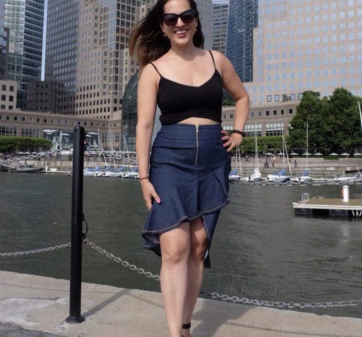 Kate standing in a black crop top and blue denim skirt in front of modern skyscrapers in lower Manhattan.