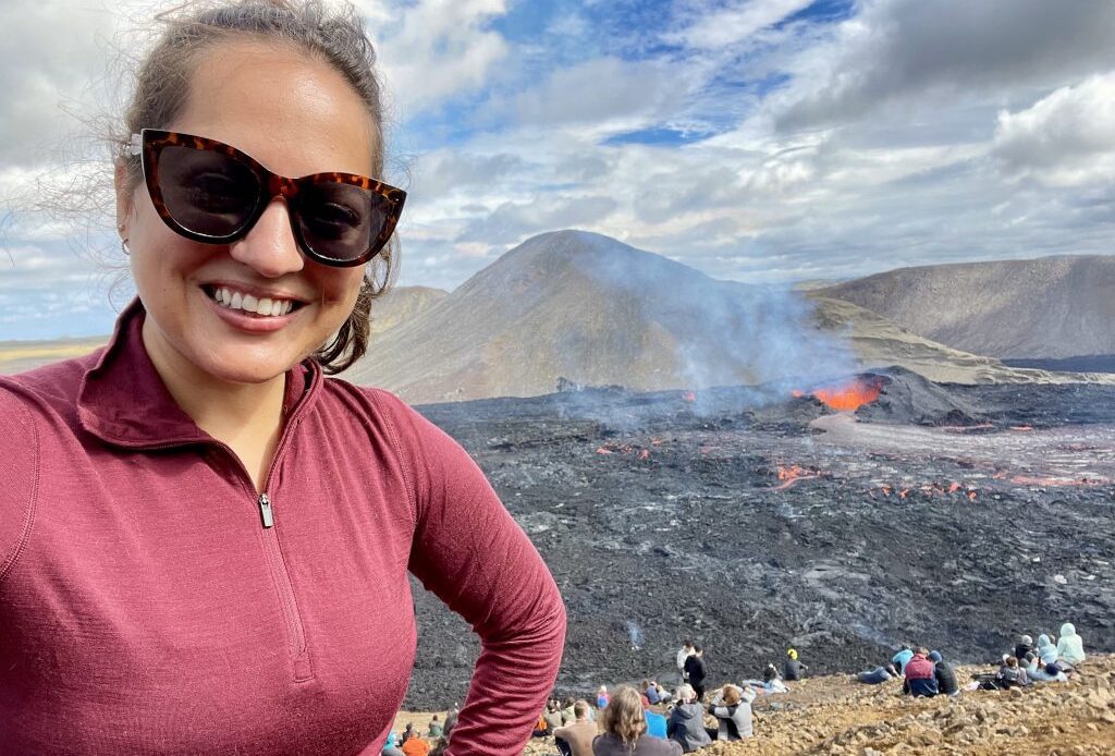 Kate takes a smiling selfie in sunglasses and a red pullover. Behind her is a smoking volcano with a crater full of bubbling red lava. You can see a bunch of people sitting on a hill watching the volcano spew.