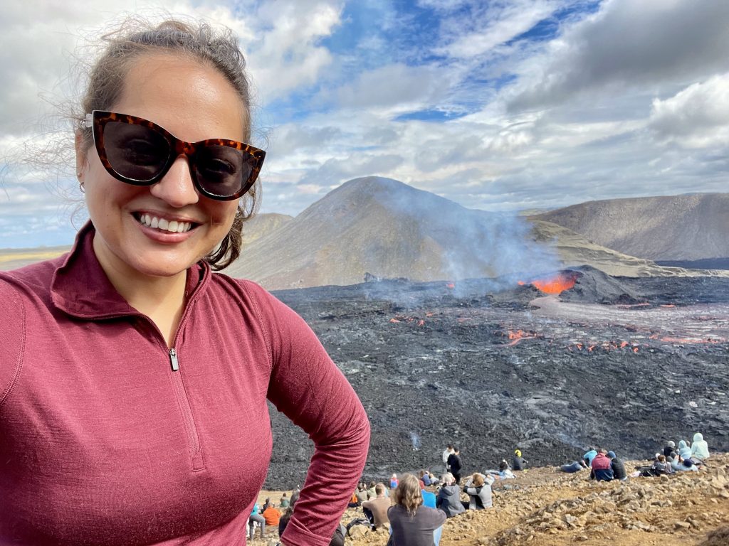 Kate takes a smiling selfie in sunglasses and a red pullover. Behind her is a smoking volcano with a crater full of bubbling red lava. You can see a bunch of people sitting on a hill watching the volcano spew.