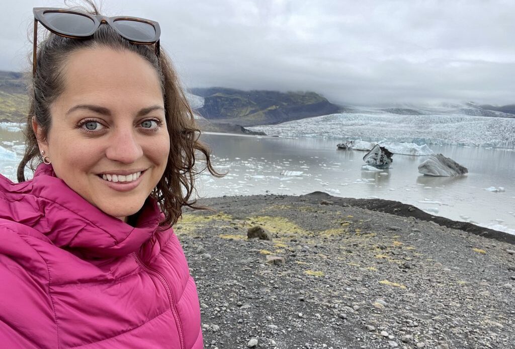 Kate wearing a pink coat and taking a smiling selfie in front of a misty lagoon filled with black-streaked icebergs, a large pale blue glacier on the mountain in the background.