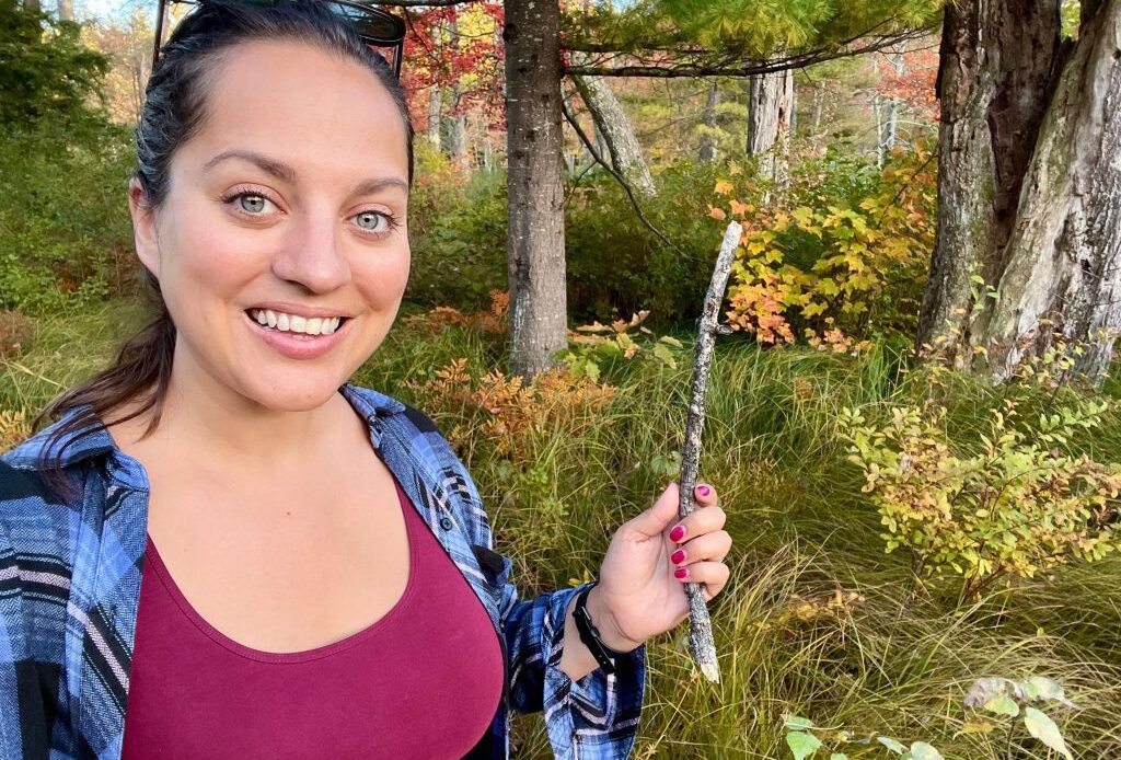 Kate wearing a blue plaid shirt over a burgundy tank top and smiling, holding up a stick in front of lots of vibrant fall foliage.