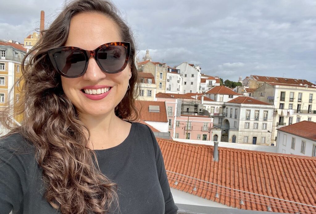 Kate takes a smiling selfie with sunglasses and long curly brown hair, the orange roofs and white buildings of hilly Lisbon behind her.