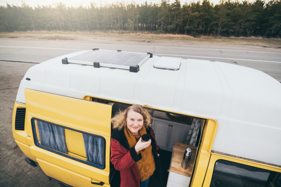 Young blond woman travelling by camper van with solar panel on the roof top and pine forest on the background