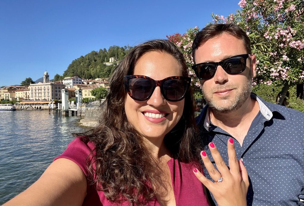 Kate and Charlie taking a smiling selfie wearing sunglasses. Kate holds up one hand and you can see an engagement ring on her finger. Behind them, Renaissance buildings set on a lake.