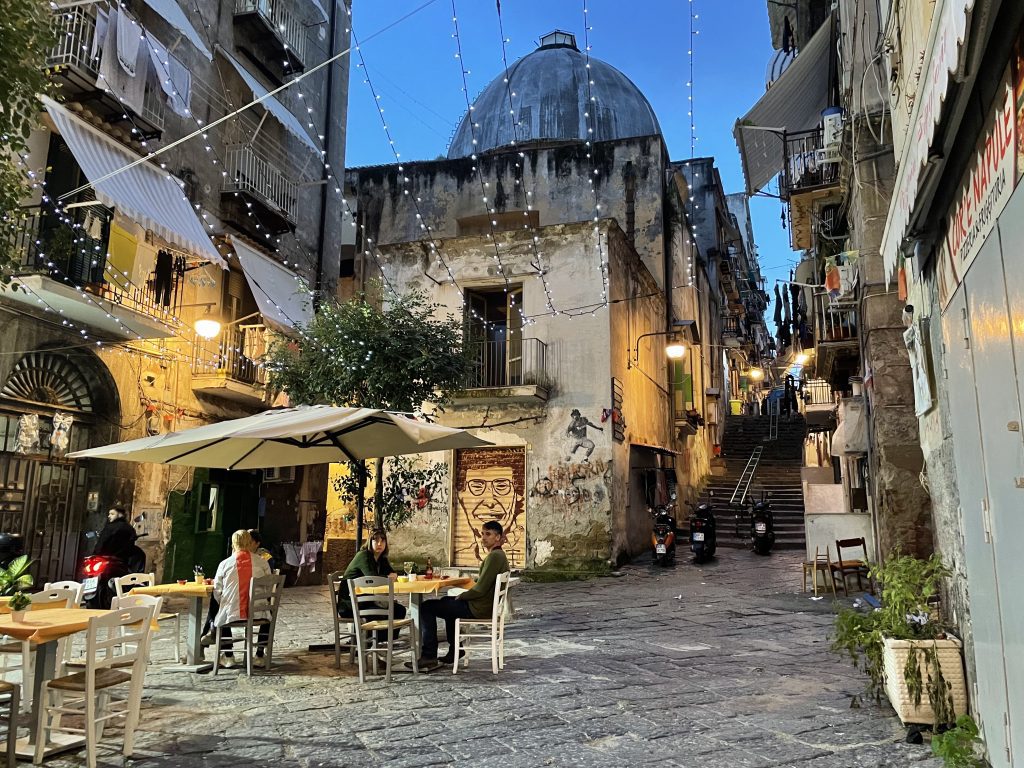 People sitting outside a cafe in an alley, as the sun begins to go down.