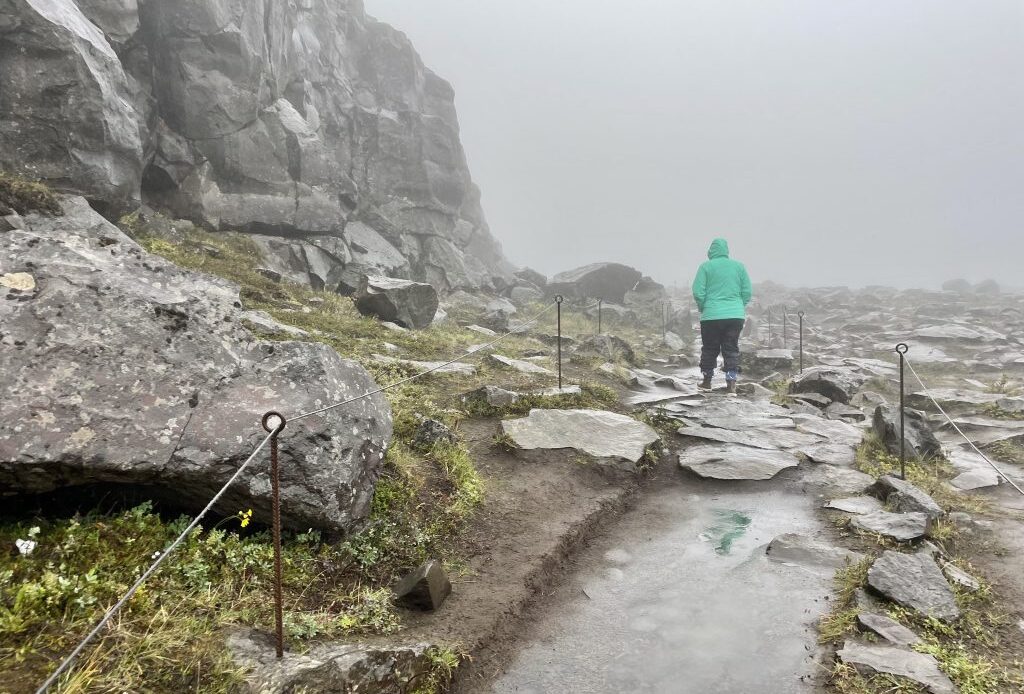 A girl in a turquoise coat walking on a rocky path in a foggy, rainy landscape.