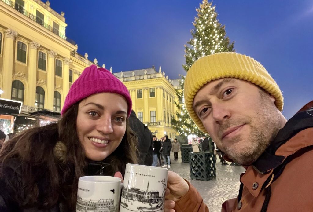 Kate and Charlie toasting gluhwein mugs with hats on in front of a bright yellow palace with a Christmas tree.