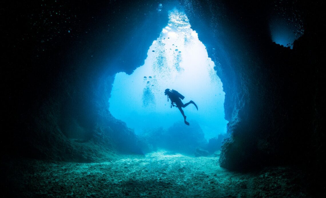 A scuba diver is in silhouette as light floods into an underwater cave