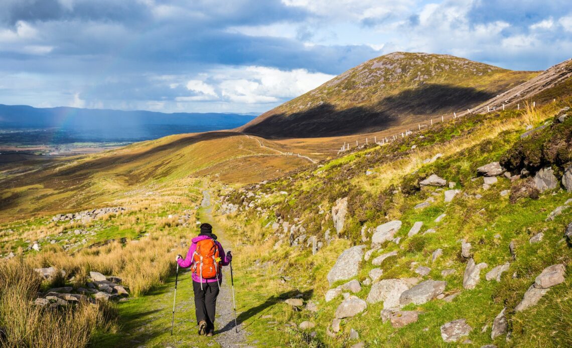 A hiker with a backpack and hiking poles follows a trail through a mountainous region of Ireland