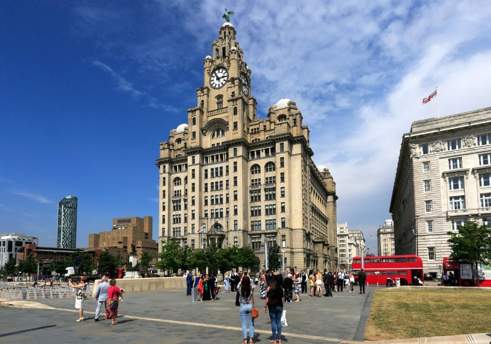 A view of the Liver Building, one of the symbols of the city of Liverpool, with a "hop on, hop off" city tour bus and some people walking around the area