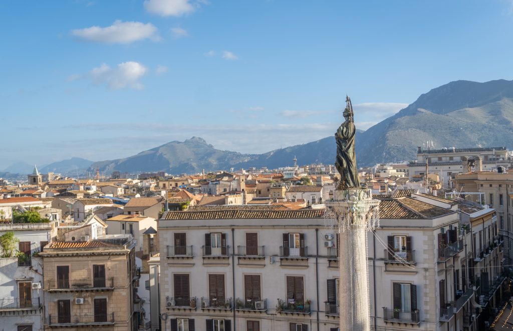 A single statue of an angel on a piazza in Palermo, in front of city roofs and mountains in the distance.