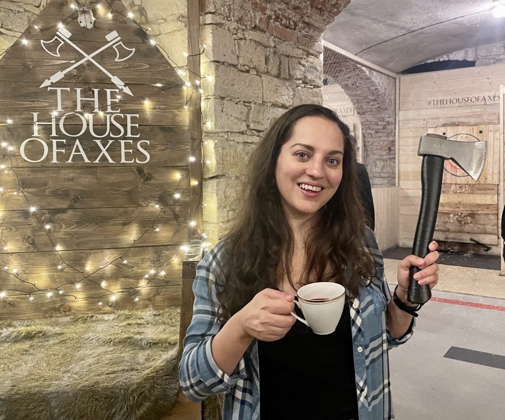Kate wearing a plaid shirt and holding a cup of tea in one hand and an axe in another in front of a sign reading "the house of axes"