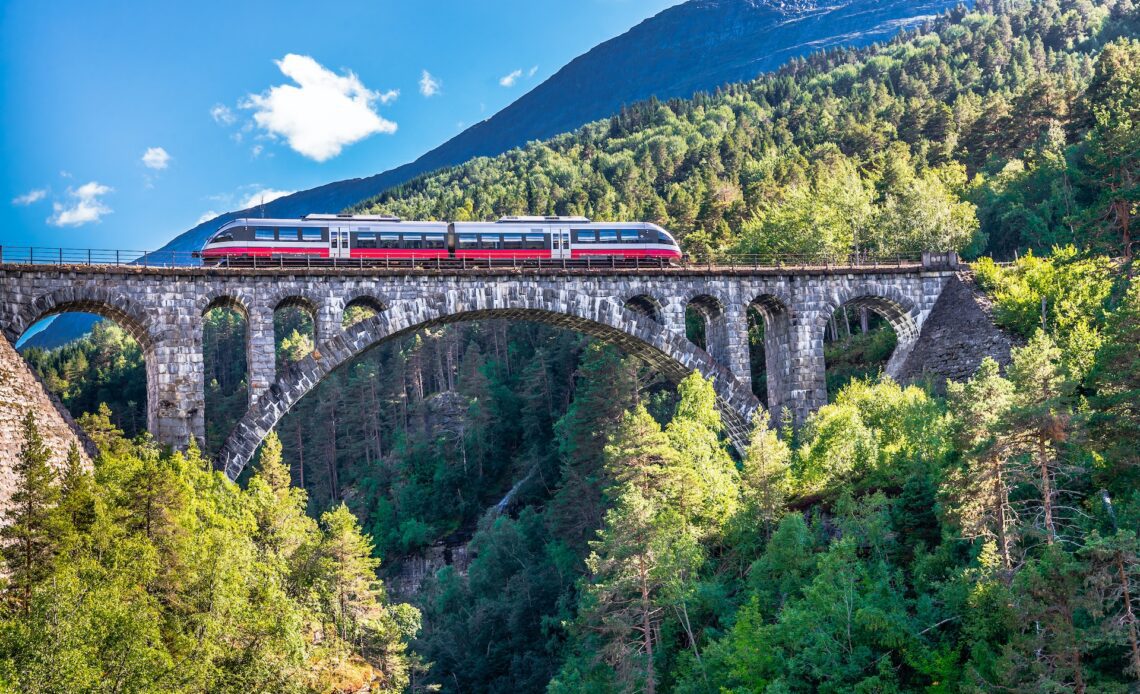 A red and white train crosses an arched railway bridge in Norway, Kylling Bridge, high above the valley tree line below with a snowy, grey mountain in the background