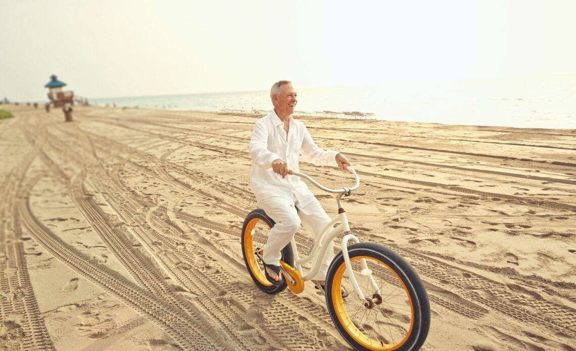 A smiling white old man with grey hair wearing all white cycles along a golden beach in Miami, US