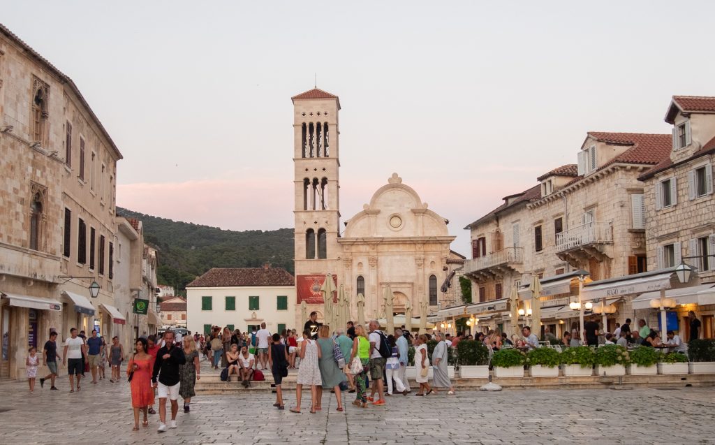 Lots of people walking in a piazza in Croatia with a white stone church with a bell tower in the center.