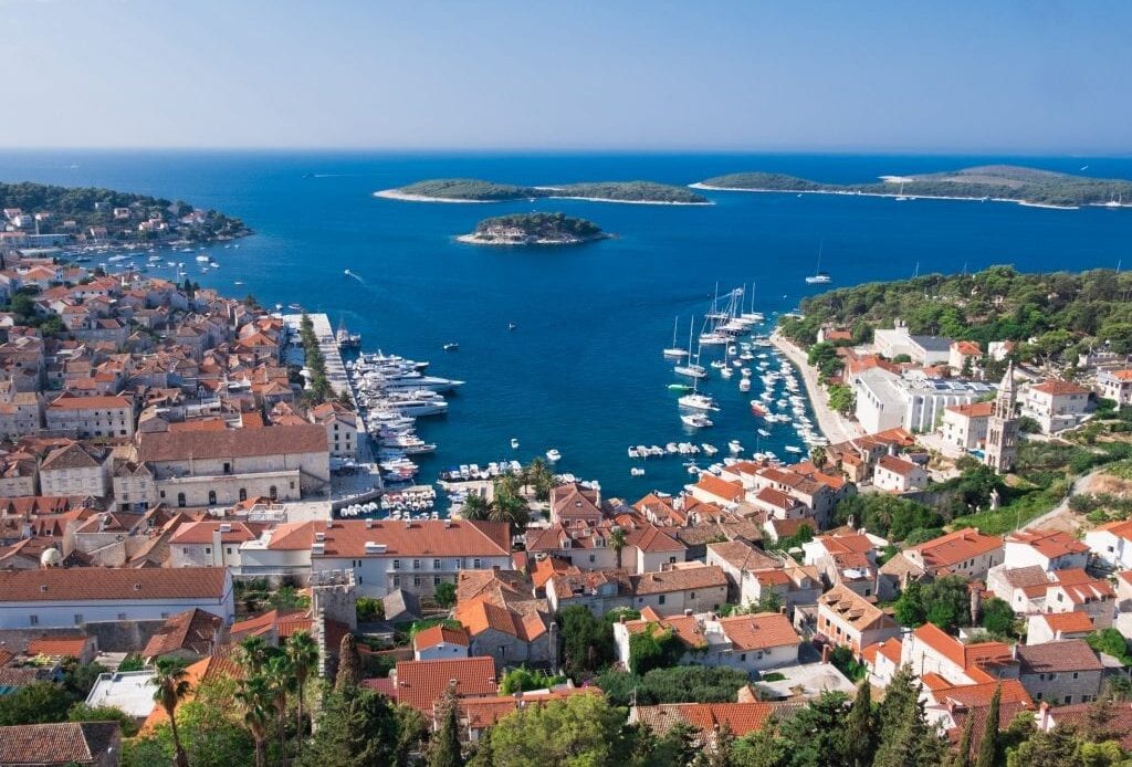 View of Hvar Town from the Spanish Fortress: a view from above, you see a small harbor surrounded with white buildings topped with orange roofs. In the distance are the green Pakleni Islands.