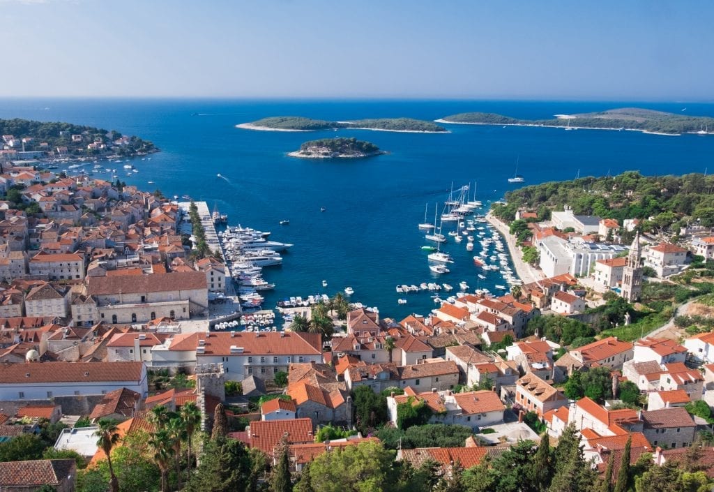 View of Hvar Town from the Spanish Fortress: a view from above, you see a small harbor surrounded with white buildings topped with orange roofs. In the distance are the green Pakleni Islands.
