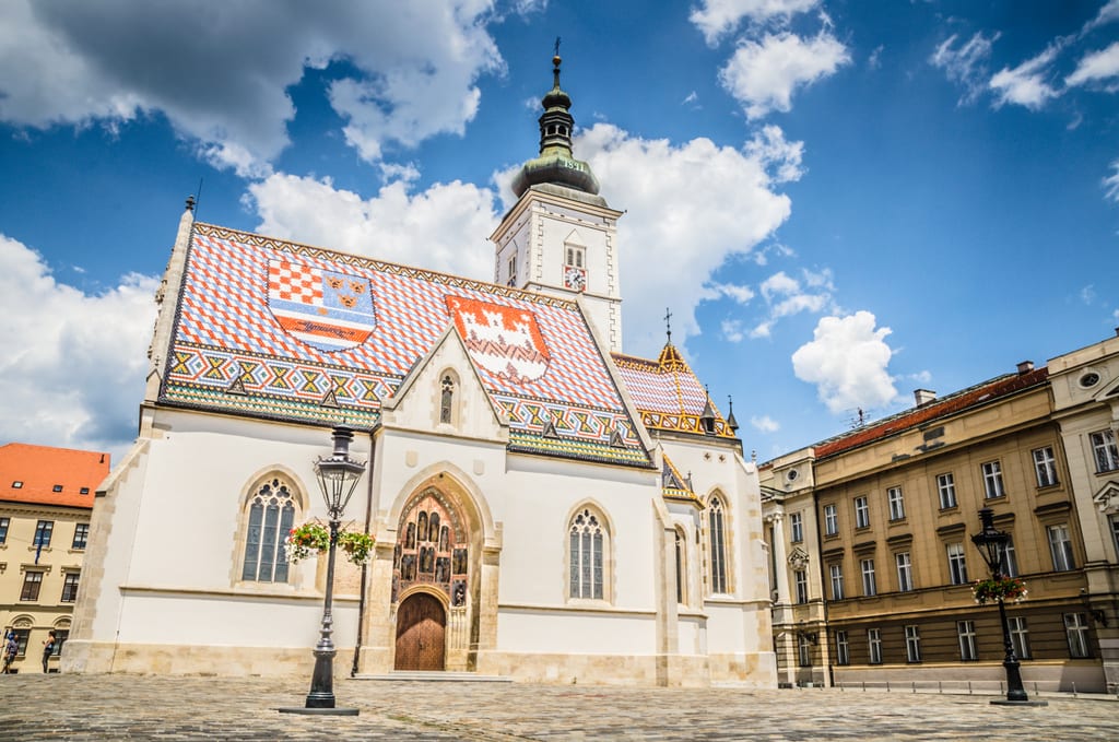 A church in Zagreb topped with orange , blue, and white tiles in a pattern underneath a bright blue sky with clouds.