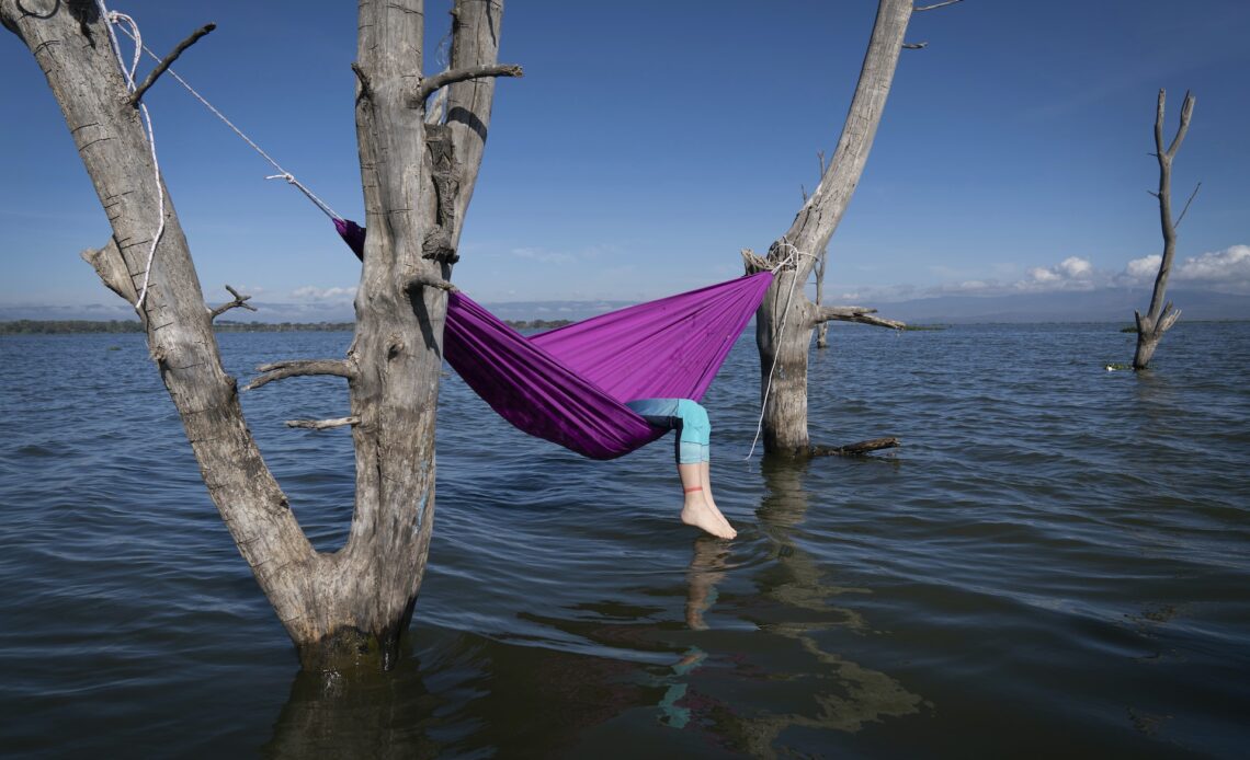 A person reclines in a hammock that is strung up between two pieces of wood over a lake