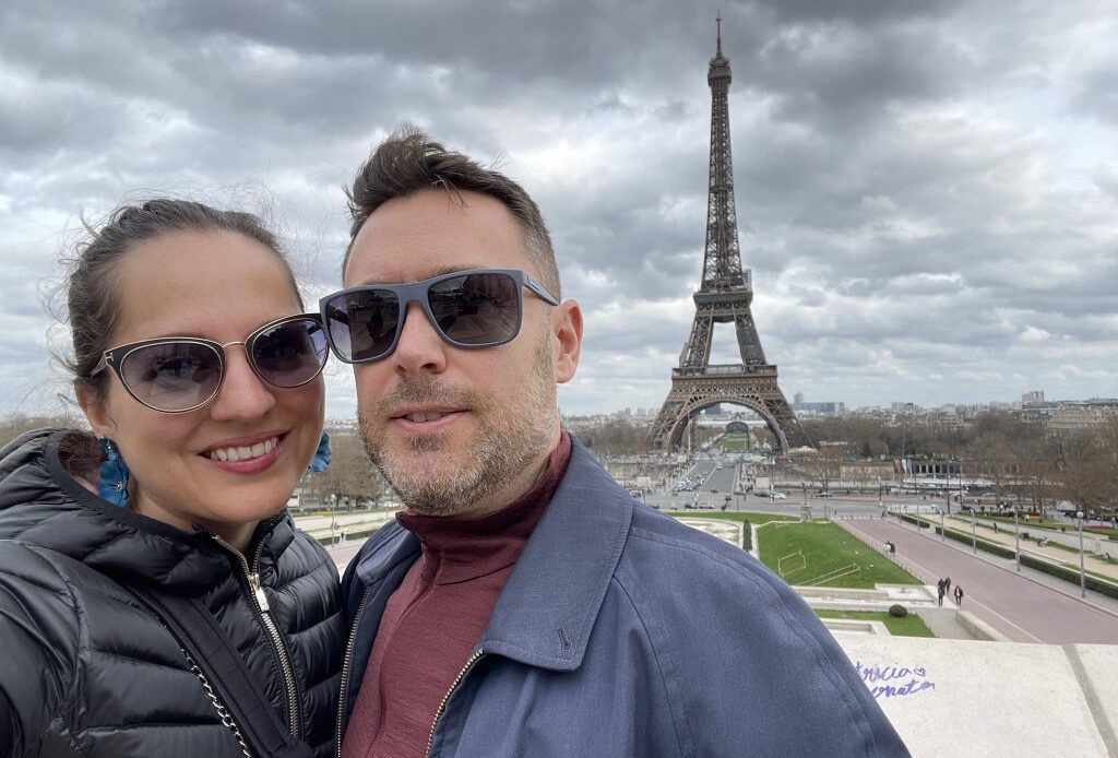 Kate and Charlie taking a selfie, wearing sunglasses, in front of the Eiffel Tower.