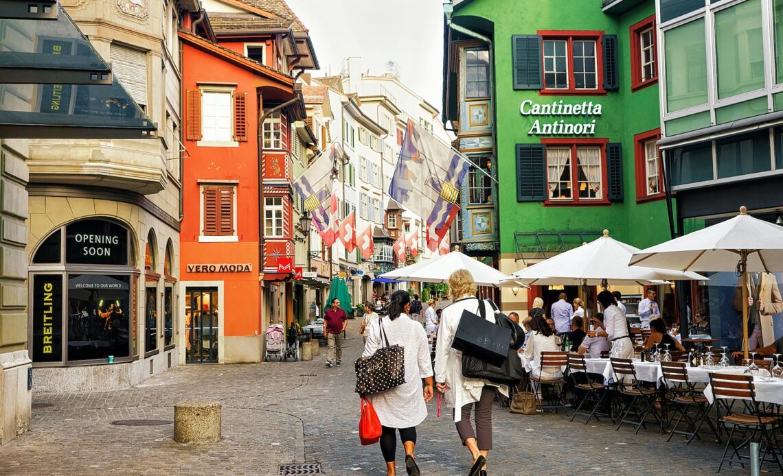 People walking down the cobbled street of the old town in Zurich, lined with colorful buildings decorated with the Swiss flag