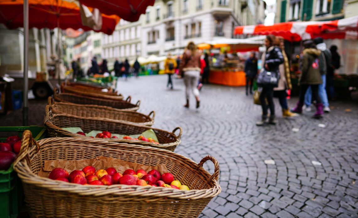 A fruit and veg stall in a cobbled city square on market day as people walk by