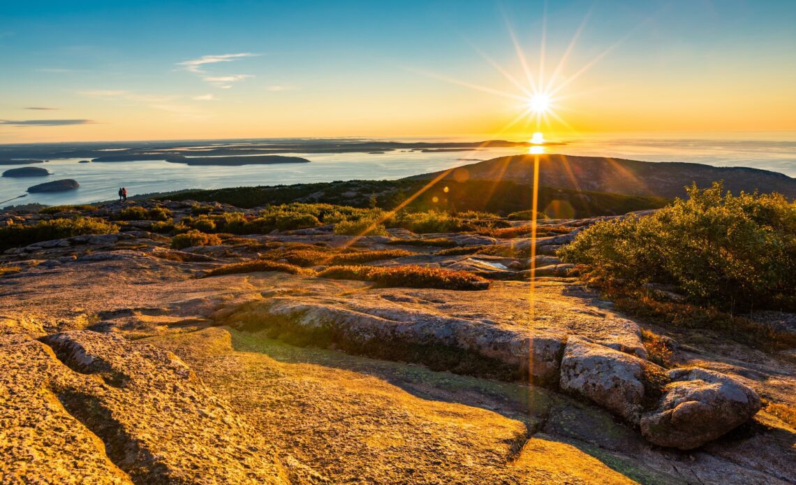 Two people walk along the edge of a mountain ridge as the sun rises in the sky casting an orange glow over the surroundings