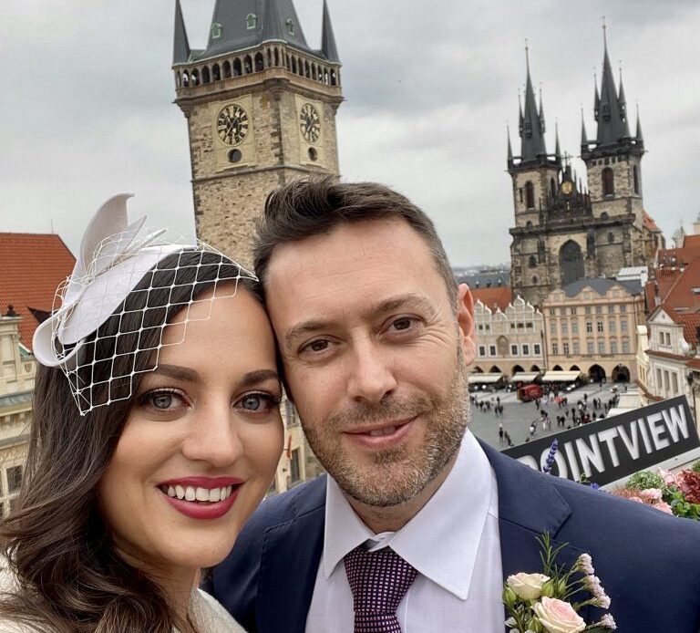 Kate and Charlie posing over the skyline view of Prague, gothic churches in the background. Kate wears a white fascinator and suit; Charlie wears a navy suit with a purple tie and a boutonniere.