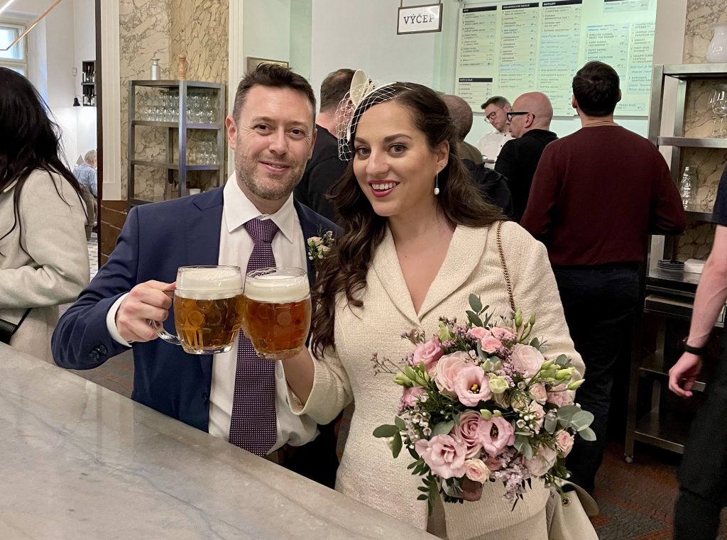 Charlie and Kate stand in a busy pub, clinking pints of beer.  They wear wedding outfits (Charlie in a navy suit and purple tie, and Kate in an ivory skirt suit and fascinator) and Kate carries a bouquet of pale pink and purple flowers.
