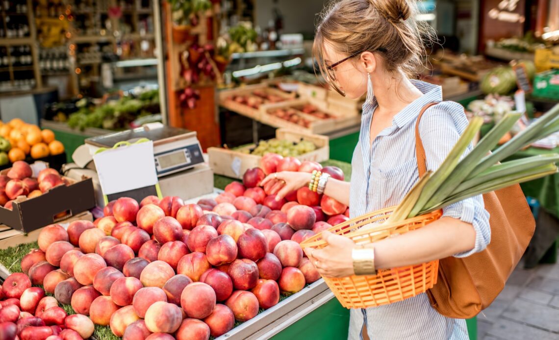 Young woman choosing a fresh peach standing with basket at an outdoor food market in France