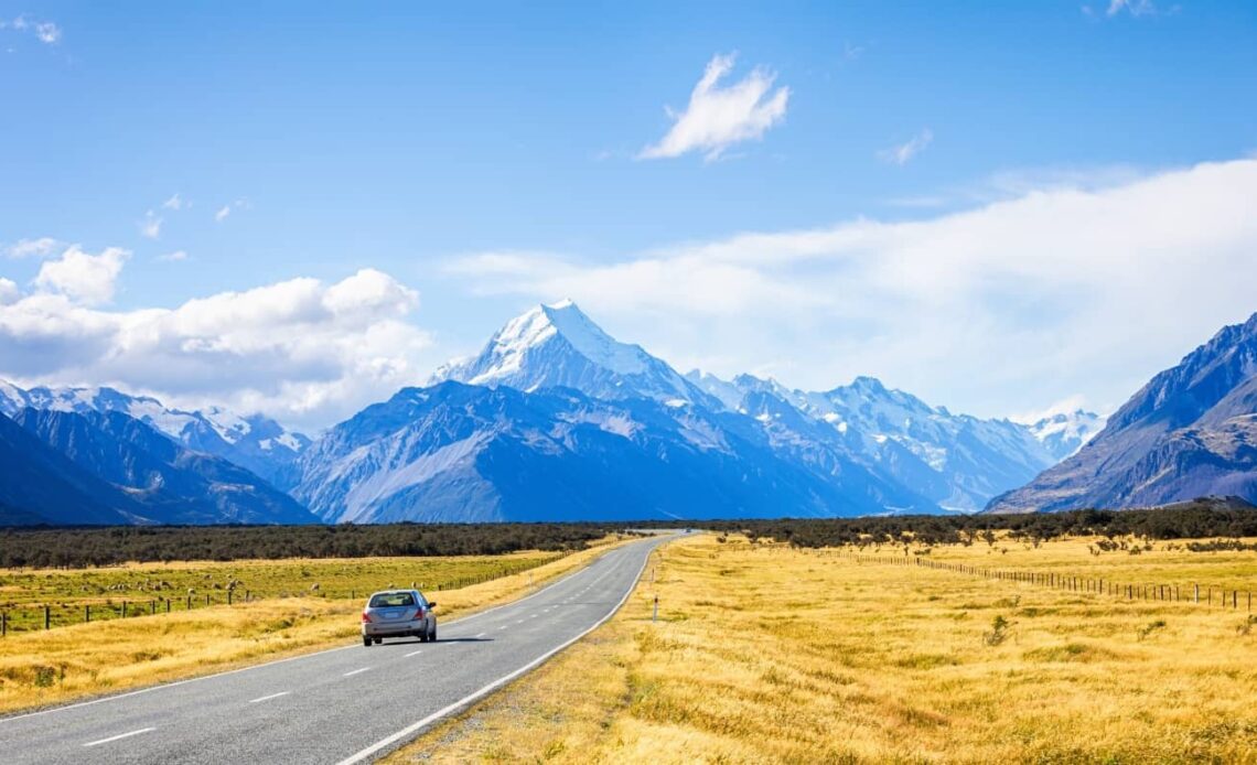 Car driving down a road with golden fields on either side, leading towards snow-capped mountains on the South Island of New Zealand
