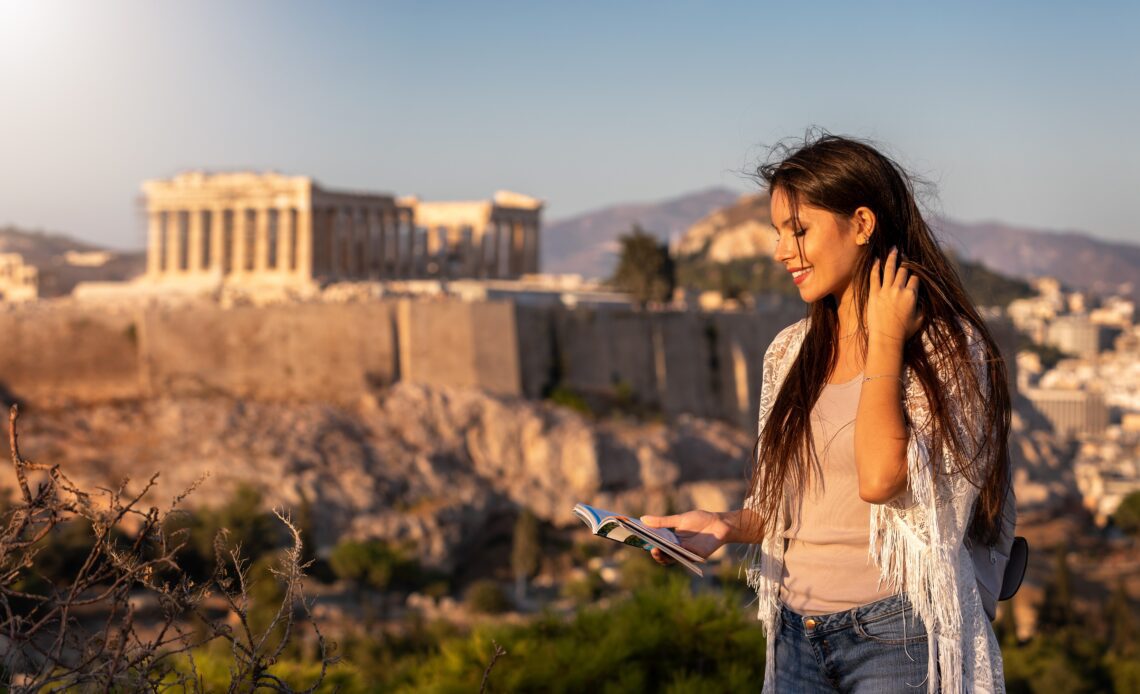 A woman with a guidebook standing in front of the Acropolis checking some facts