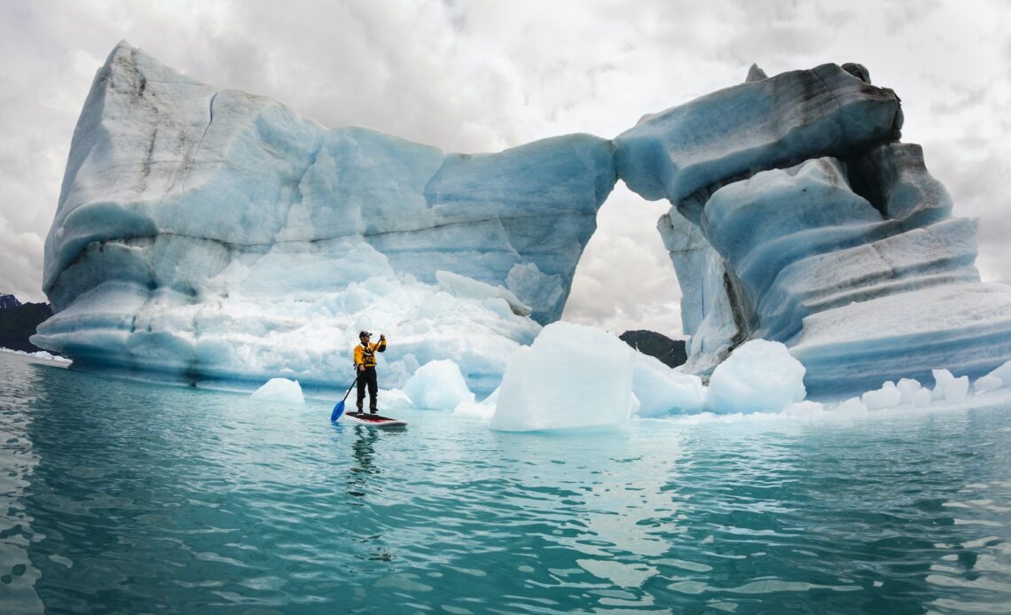 A solo figure on a stand up paddle board (SUP) paddles past icebergs