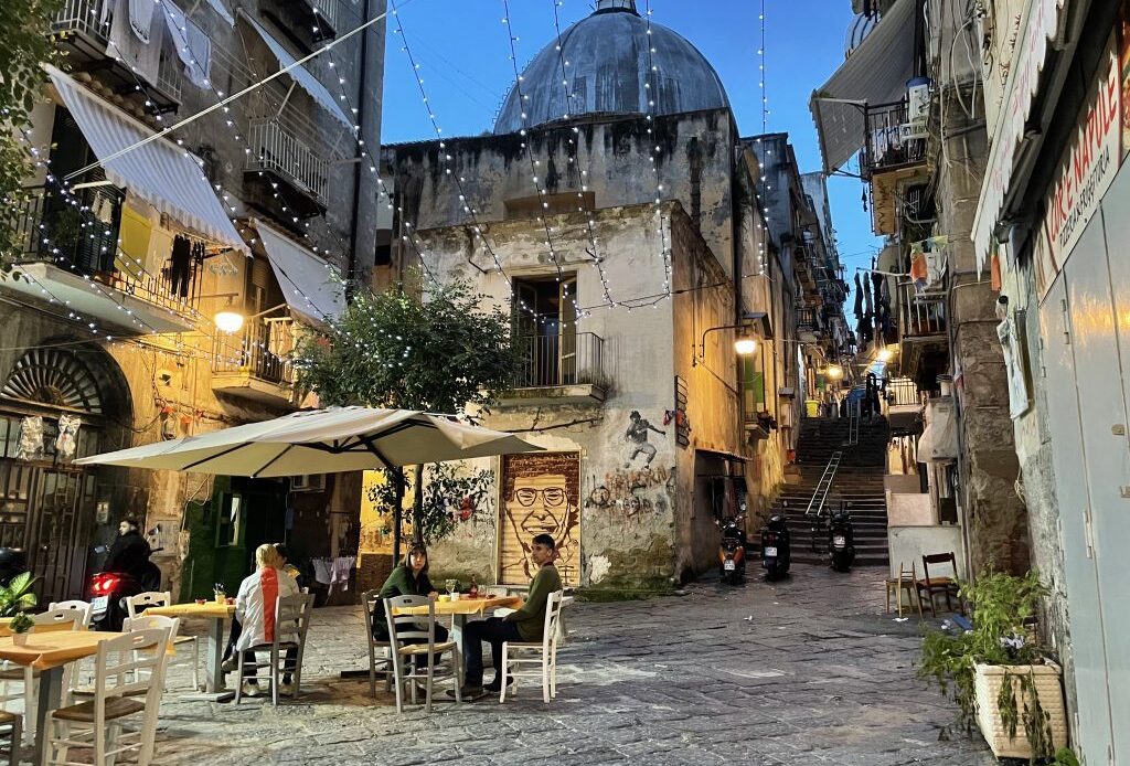 People sitting outside a cafe in an alley, as the sun begins to go down.