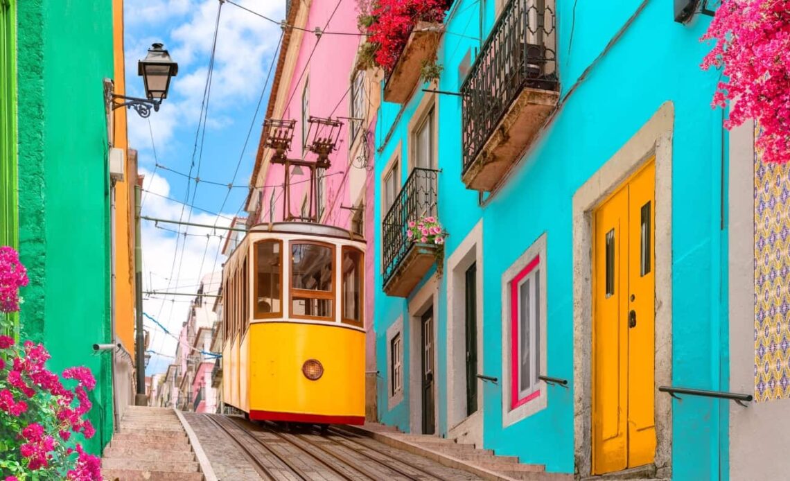 Historic yellow tram on a street going down a hill lined with colorful buildings and flowers on the balconies in Lisbon, Portugal