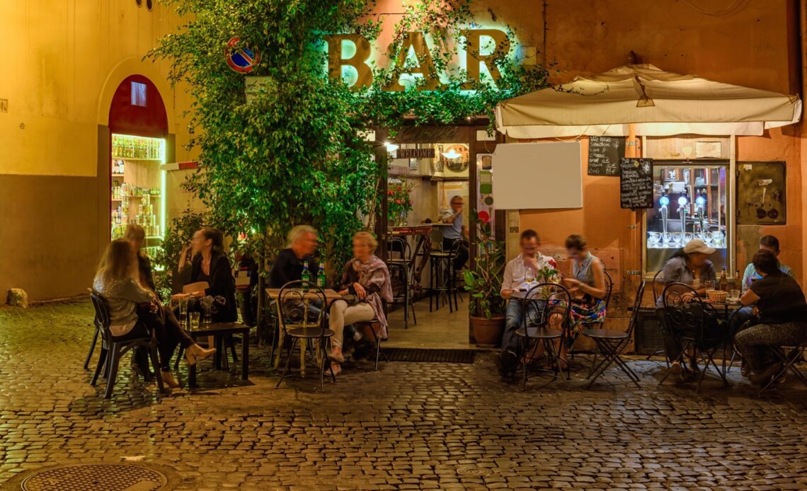 People sit outside a bar on an old street at night in Trastevere, Rome