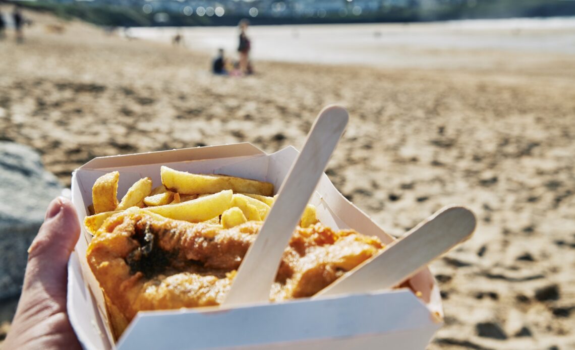 A hand holding a box of fish and chips at the beach on a bright sunny day