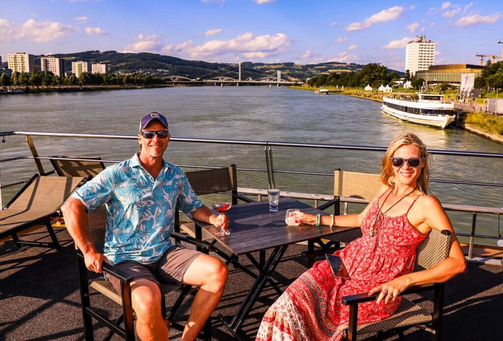 Man and lady having drinks on the deck of a cruise ship with a river behind them