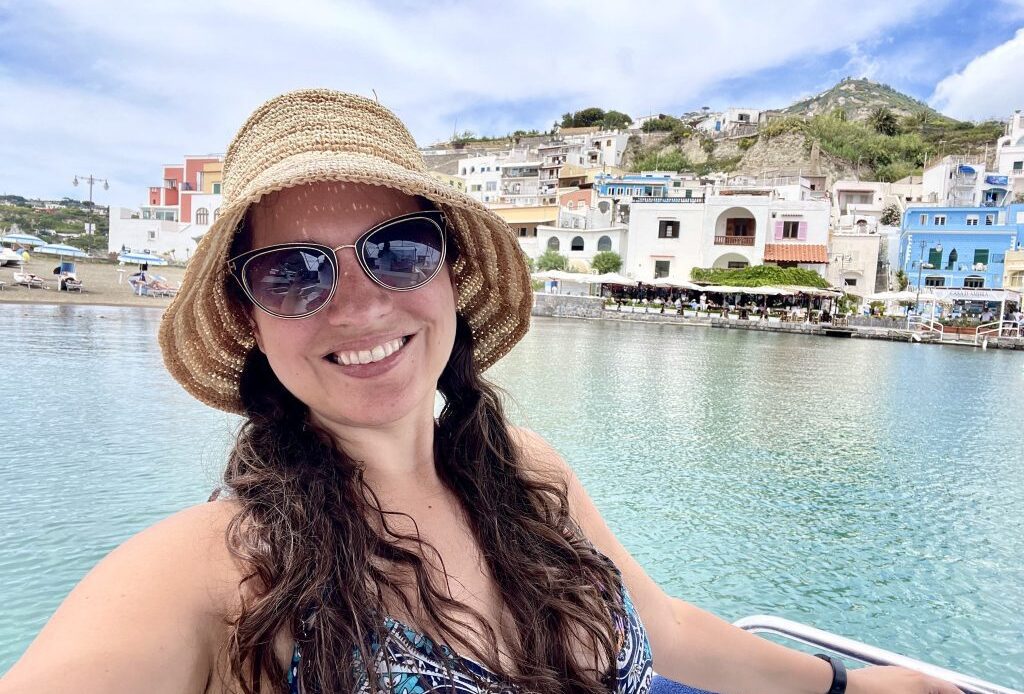 Kate smiling, wearing a straw bucket hat and sunglasses, in front of a small coastal town in Ischia, Italy.