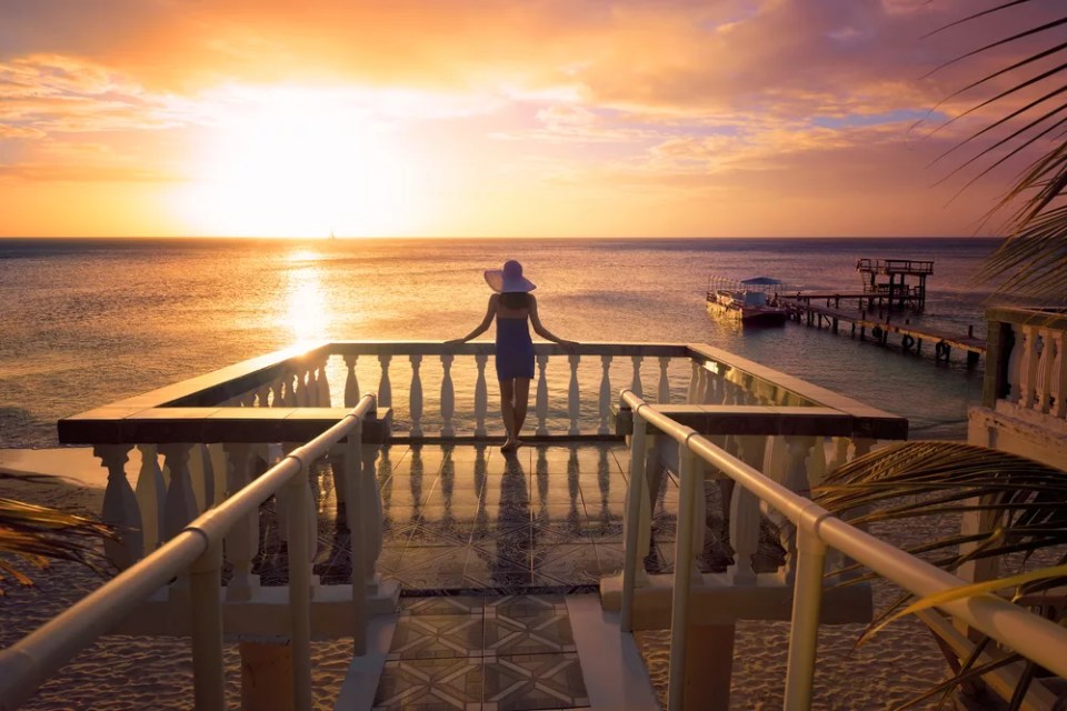 A woman in a hat looking at the romantic Caribbean sunset while standing on the balcony. Roatan Island, Honduras